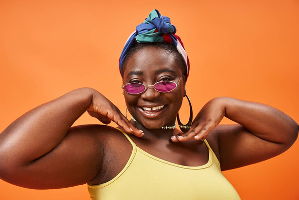 lichaam positief, gelukkig plus size Afrikaans amerikaanse vrouw in hoofddoek en trendy zonnebril op oranje - Foto, afbeelding