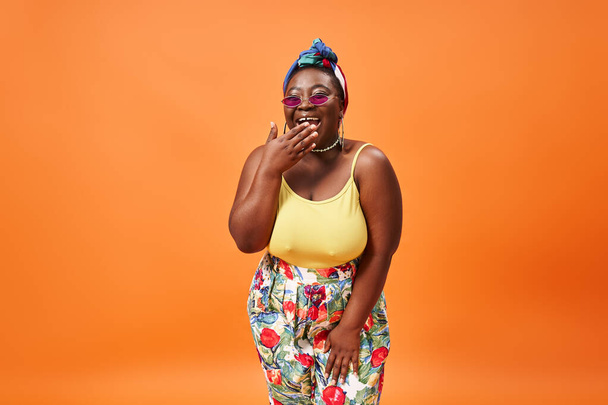 szczęśliwy plus rozmiar Afroamerykanka kobieta w kwiatowym stroju i okulary uśmiecha się na pomarańczowym tle - Zdjęcie, obraz