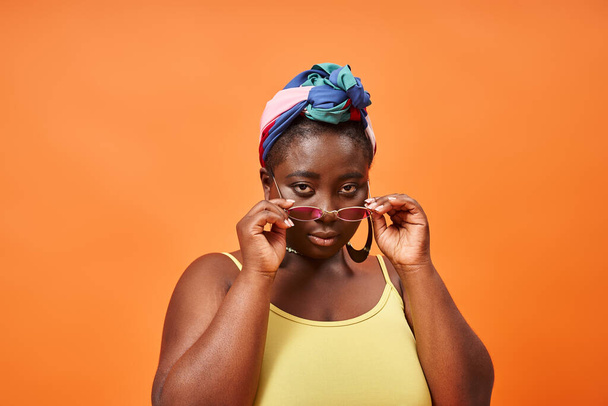 młody plus rozmiar afrykański amerykański kobieta w chusta i modne okulary przeciwsłoneczne na pomarańczowym tle - Zdjęcie, obraz