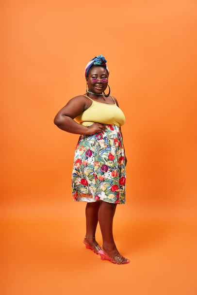 plusz méret afro-amerikai modell virágos ruhában és napszemüveg pózol kézzel csípő narancs - Fotó, kép