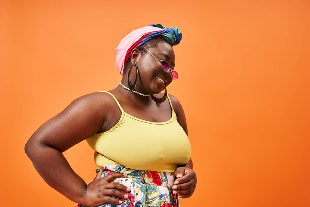 Artı, çiçekli kıyafetli, güneş gözlüklü, eli turuncunun üstünde poz veren Afrikalı Amerikalı kadın. - Fotoğraf, Görsel
