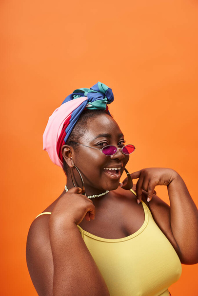 radosne plus size afrykański amerykański kobieta w chusta i stylowe okulary przeciwsłoneczne na pomarańczowym tle - Zdjęcie, obraz