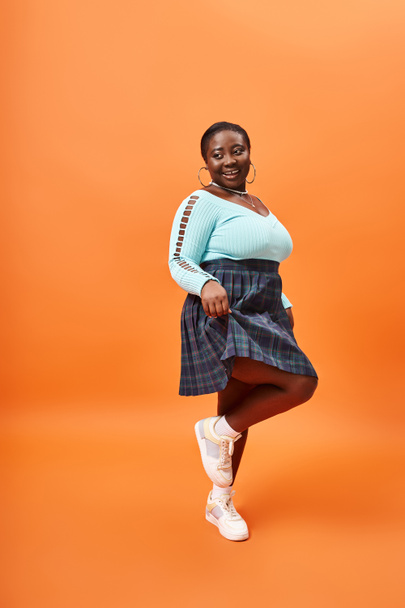 happy plus size Afrykańska kobieta w kratę spódnica i niebieski długi rękaw na pomarańczowym tle - Zdjęcie, obraz