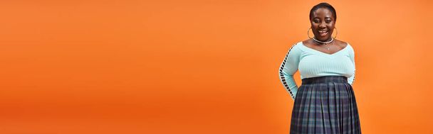 Körper positive Afroamerikanerin in kariertem Rock und blauem Langarm lächelnd auf orangefarbenem Banner - Foto, Bild