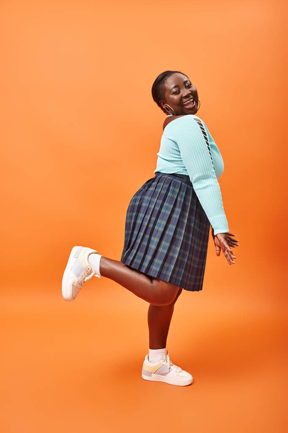 playful plus size african american model in plaid skirt and mint color jumper posing on orange - Photo, Image