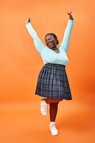 alegre modelo afroamericano de talla grande en falda a cuadros y jersey de color menta posando en naranja - Foto, imagen
