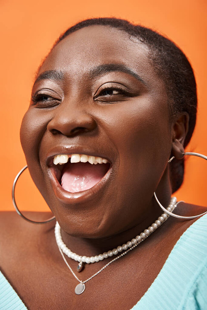 portrait de femme afro-américaine rayonnante plus la taille en bleu manches longues riant sur orange - Photo, image