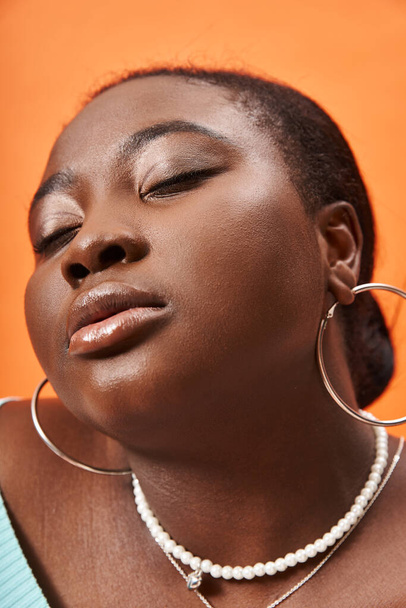 portrét zamyšlené plus velikosti africké americké ženy pózující se zavřenýma očima na oranžovém pozadí - Fotografie, Obrázek
