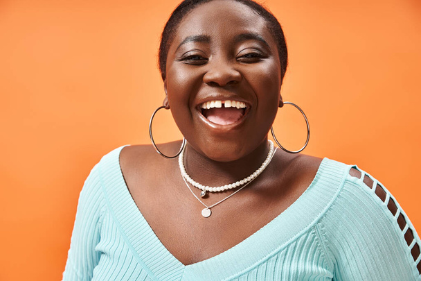 retrato de alegre más tamaño mujer afroamericana en azul manga larga riendo en naranja - Foto, Imagen