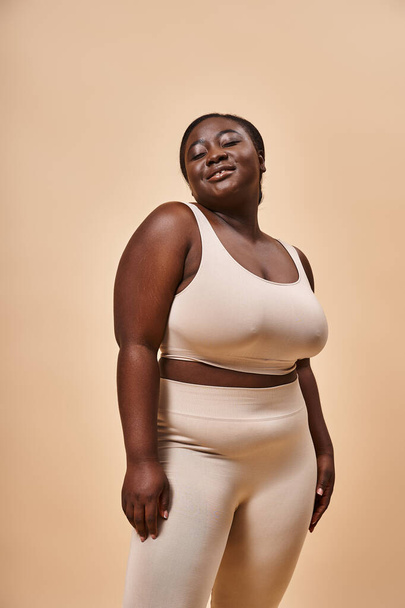 Stralende plus size Afrikaanse Amerikaanse vrouw in beige sportkleding glimlachen op bijpassende achtergrond - Foto, afbeelding