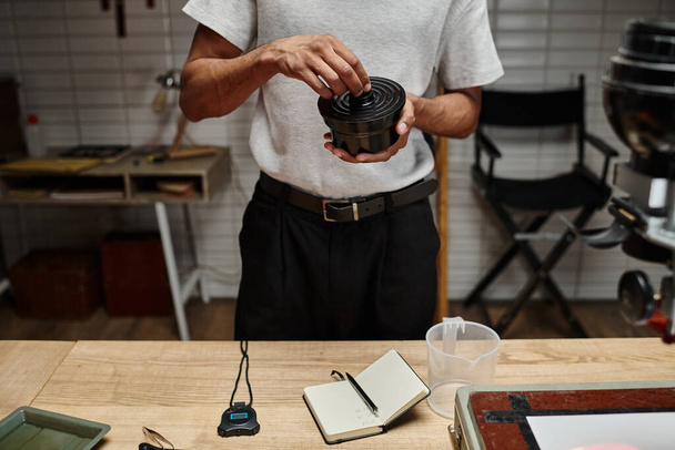 homem afro-americano cultivado segurando recipiente de filme na câmara escura, passo no processo de desenvolvimento analógico - Foto, Imagem