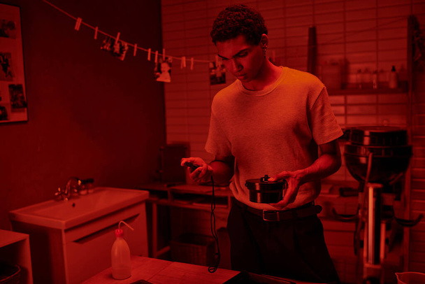 φωτογράφος σε κόκκινο-φωτισμένο δωμάτιο, μαύρος άνθρωπος χειρίζεται προσεκτικά την ανάπτυξη ταινιών με χρονοδιακόπτη σκοτεινό θάλαμο - Φωτογραφία, εικόνα