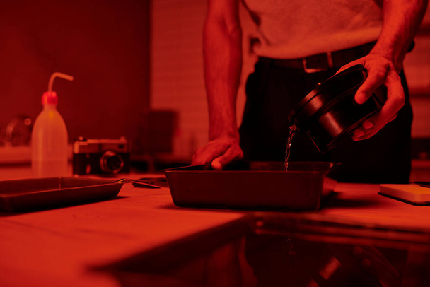 visão recortada do fotógrafo derrama desenvolvedor em bandeja para processamento de filmes em câmara escura vermelha - Foto, Imagem