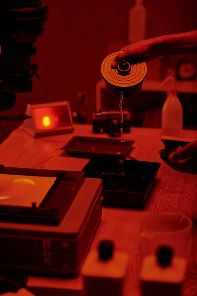 vista recortada de fotógrafo vierte líquido desarrollador para el procesamiento de películas, cuarto oscuro rojo - Foto, imagen