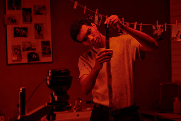 Odaklanmış Afrikalı Amerikalı fotoğrafçı film şeridi geliştirdi, kırmızı karanlık odada yıkandı. - Fotoğraf, Görsel
