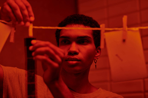 fotógrafo afroamericano cuelga tira de película recién desarrollada en un cuarto oscuro con luz roja, atemporal - Foto, imagen