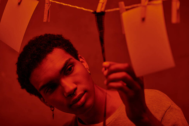 Genç Afrikalı Amerikalı adam kırmızı ışıklı karanlık bir odada yeni geliştirilmiş bir film şeridine bakıyor. - Fotoğraf, Görsel