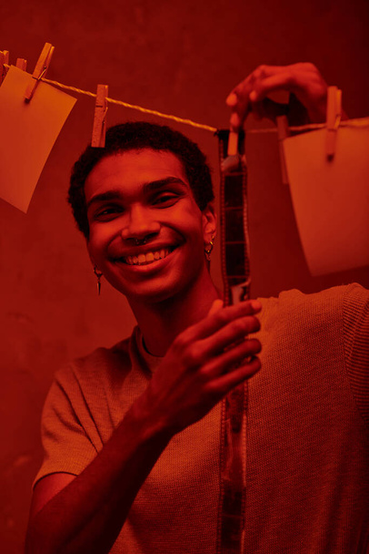 fröhlicher afrikanisch-amerikanischer Mann, der frisch entwickelte Filmstreifen in einer rotbeleuchteten Dunkelkammer hängt, Nostalgie - Foto, Bild