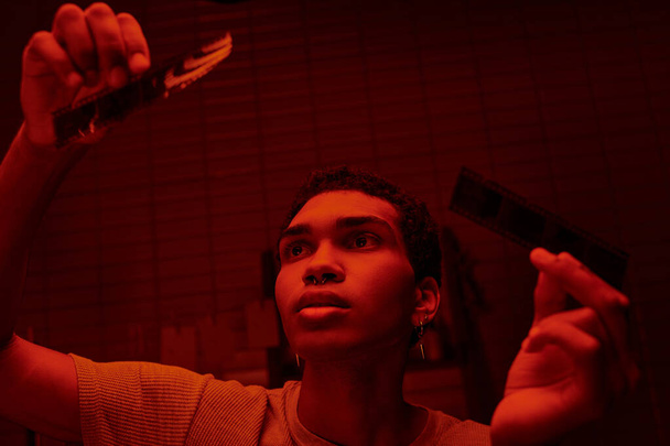 fotógrafo afroamericano enfocado comparando tiras de película desarrolladas en un cuarto oscuro iluminado por el rojo - Foto, Imagen