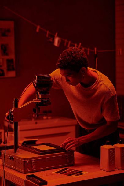アフリカ系アメリカ人の写真家が暗室の赤い安全ライトの下でネガティブな写真を検査 - 写真・画像