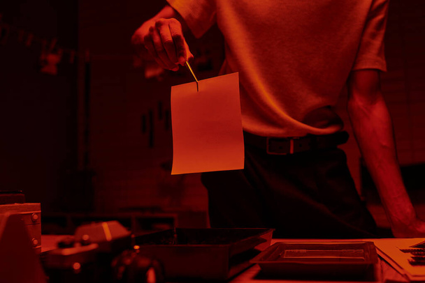 φωτογραφία φωτογράφου να κρατάει τσιμπιδάκι με φωτογραφικό χαρτί σε σκοτεινό θάλαμο με κόκκινο φως - Φωτογραφία, εικόνα
