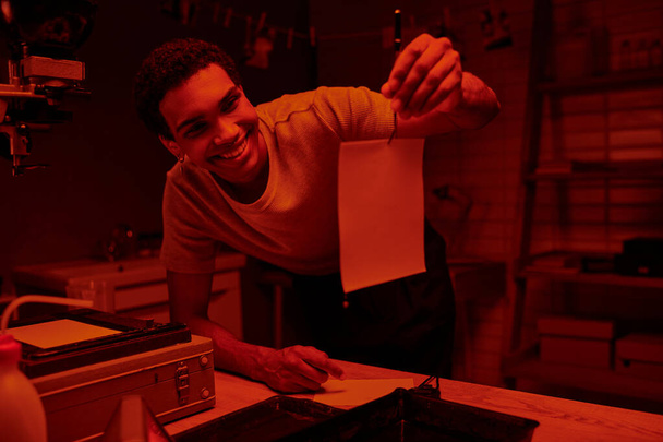 щасливий афроамериканський фотограф тримає пінцет із свіжорозробленим фотопапером у темній кімнаті - Фото, зображення