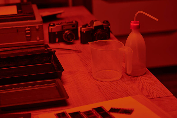 Een tafel met analoge camera en maatbeker voor filmontwikkeling in donkere kamer met rood licht - Foto, afbeelding