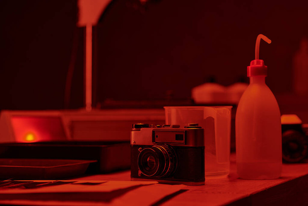 τραπέζι με αναλογική κάμερα και διάφορα εργαλεία για την ανάπτυξη ταινιών σε σκοτεινό θάλαμο με κόκκινο φως - Φωτογραφία, εικόνα