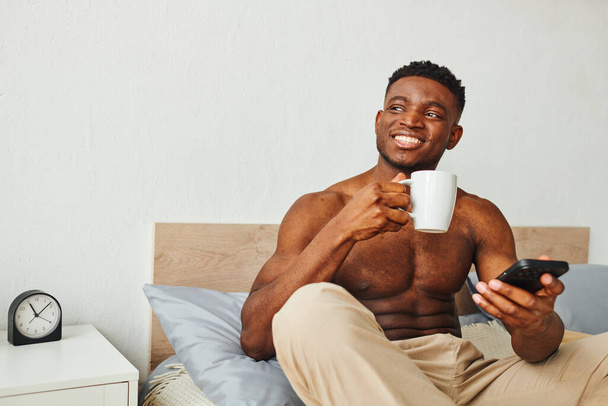 щасливий без сорочки афроамериканський чоловік з чашкою кави та смартфоном, сидячи та дивлячись на ліжко - Фото, зображення