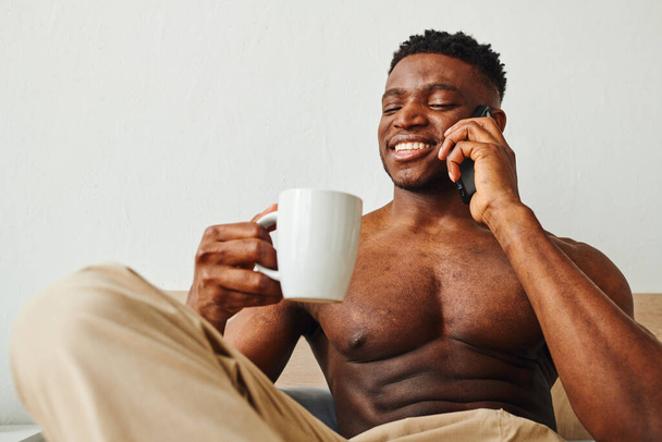 alegre muscular africano americano homem com café da manhã falando no smartphone no quarto - Foto, Imagem