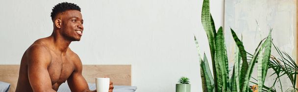 sonriente afroamericano sin camisa hombre sentado con taza de café cerca de plantas verdes en el dormitorio, pancarta - Foto, Imagen