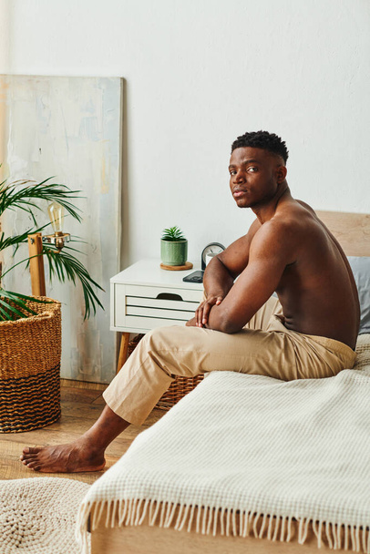 doordachte Afrikaans-Amerikaanse shirtloze man in pyjamabroek zittend en kijkend naar camera in slaapkamer - Foto, afbeelding