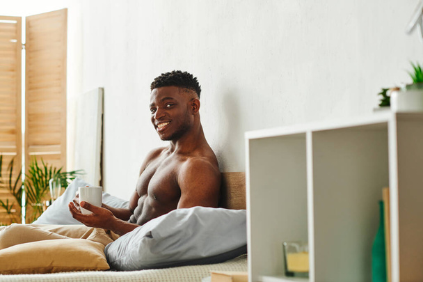 gelukkig Afrikaans amerikaanse man met gespierd lichaam zitten met koffiekop kijken naar camera in slaapkamer - Foto, afbeelding