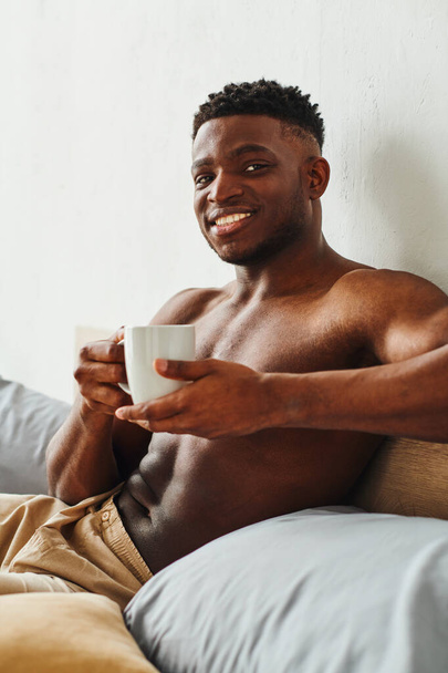 glimlachende Afro-Amerikaanse man met shirtloze romp met koffiebeker en kijkend naar camera op bed - Foto, afbeelding