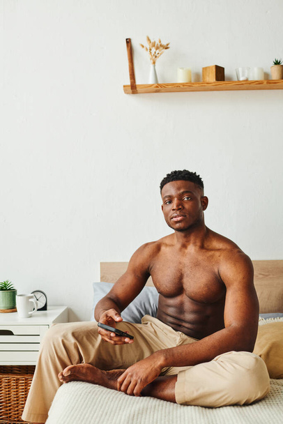 svalnatý africký Američan v pyžamových kalhotách sedí na posteli s smartphonem a dívá se do kamery - Fotografie, Obrázek