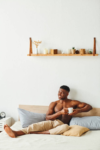 χαρούμενος αθλητικός Αφροαμερικάνος άνδρας με παντελόνι πιτζάμας κάθεται με φλιτζάνι καφέ στο κρεβάτι το πρωί - Φωτογραφία, εικόνα