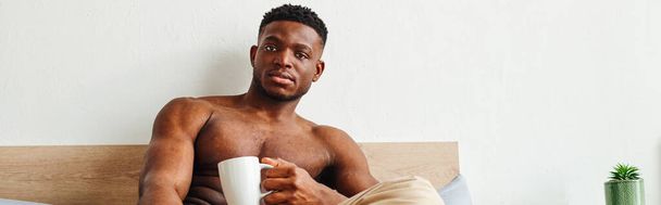 sterke en knappe Afro-Amerikaanse man met koffiekopje kijken naar camera in de slaapkamer, banner - Foto, afbeelding