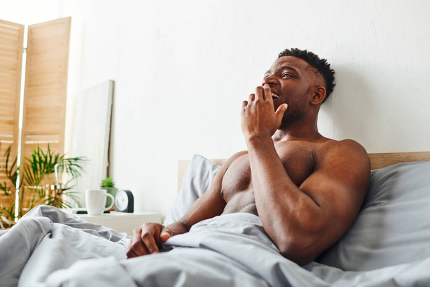 glücklich hemdlos afrikanisch-amerikanischer Mann mit starkem Körper, der morgens aufwacht und auf dem Bett gähnt - Foto, Bild