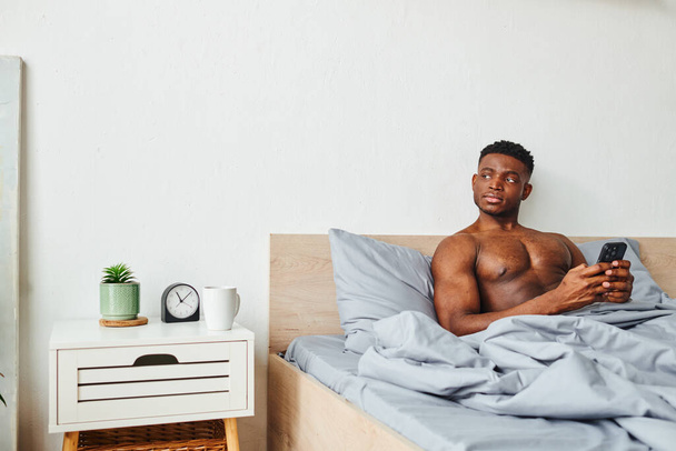 przemyślany Afrykański amerykański mężczyzna z bezkoszulkowym muskularnym ciałem i smartfonem odwracając wzrok w sypialni - Zdjęcie, obraz