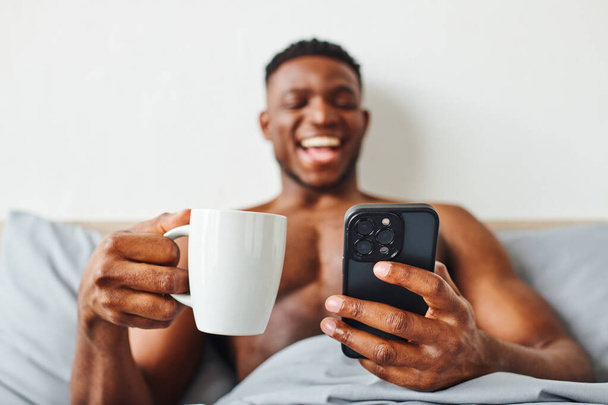opgewonden shirtloze Afrikaanse Amerikaanse man met 's morgens koffie en mobiele telefoon lachen in de slaapkamer - Foto, afbeelding
