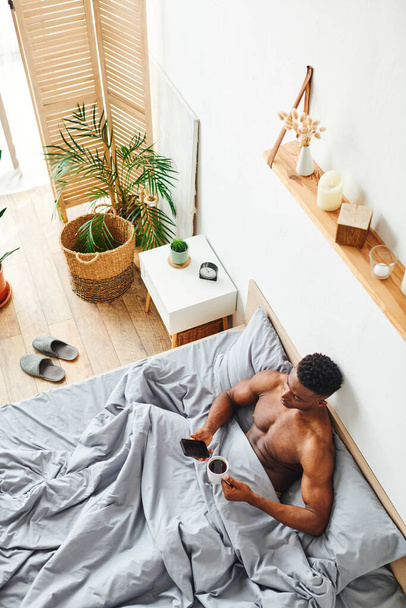 top view of handsome african american man with morning coffee and smartphone on cozy bed at home - Photo, Image