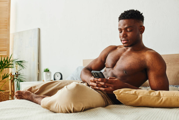 atlético afroamericano hombre en pantalones de pijama mensajería en el teléfono móvil en la cama acogedora por la mañana - Foto, Imagen