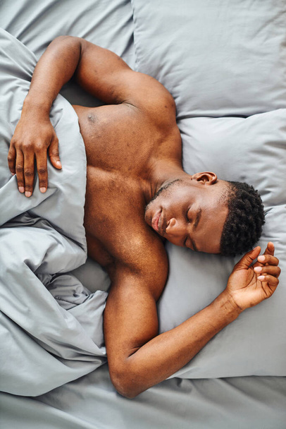 κορυφαία άποψη του νεαρού Αφροαμερικανού άντρα που κοιμάται σε άνετο κρεβάτι κάτω από μαλακή κουβέρτα το πρωί - Φωτογραφία, εικόνα
