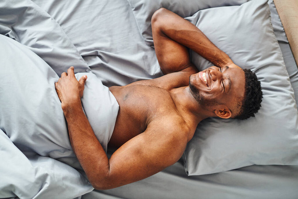 bovenaanzicht van gelukkig gespierd Afrikaans amerikaanse man dromen en glimlachen op grijs comfortabel beddengoed - Foto, afbeelding