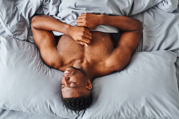 vista dall'alto dell'uomo afro-americano con un corpo forte che sogna su lenzuola comode grigie a casa - Foto, immagini
