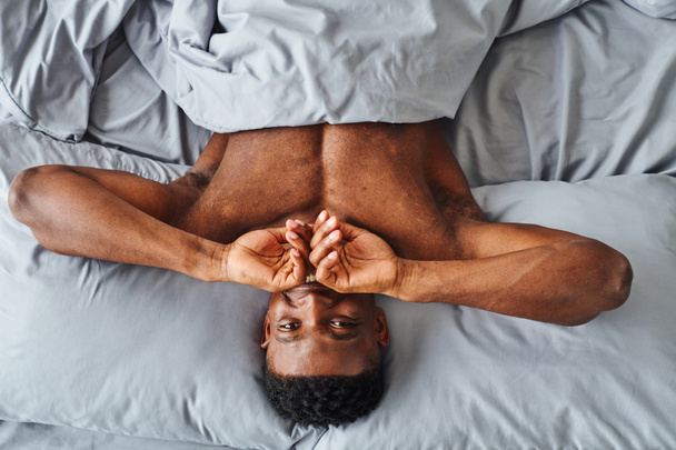 Вид сверху жизнерадостного африканского мужчины, закрывающего рот и смотрящего на камеру, просыпающегося на кровати - Фото, изображение