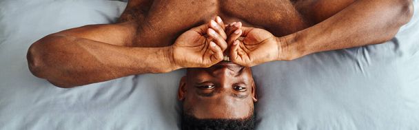 radosny Afrykańczyk zakrywający usta i uśmiechający się do aparatu budzącego się na łóżku, widok z góry, baner - Zdjęcie, obraz