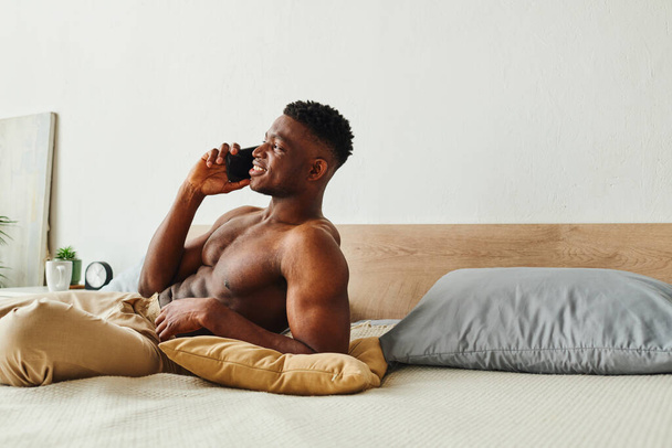 szczęśliwy Afrykanin bez koszuli amerykański człowiek w piżamie spodnie rozmowy na smartfonie w pobliżu poduszki na przytulnym łóżku - Zdjęcie, obraz