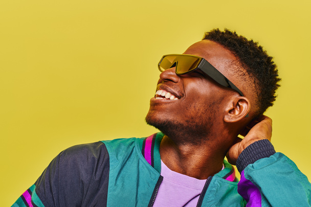 despreocupado hombre afroamericano en chaqueta cortavientos y gafas de sol con estilo mirando hacia otro lado en amarillo - Foto, imagen