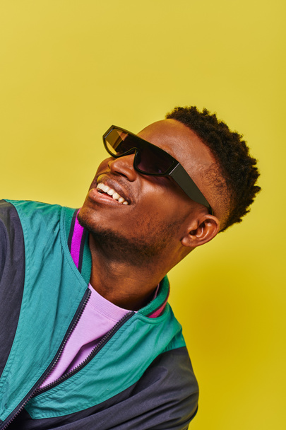 portrét šťastného afrického Američana v slunečních brýlích a světlé bundě odvracející pohled na žlutou - Fotografie, Obrázek